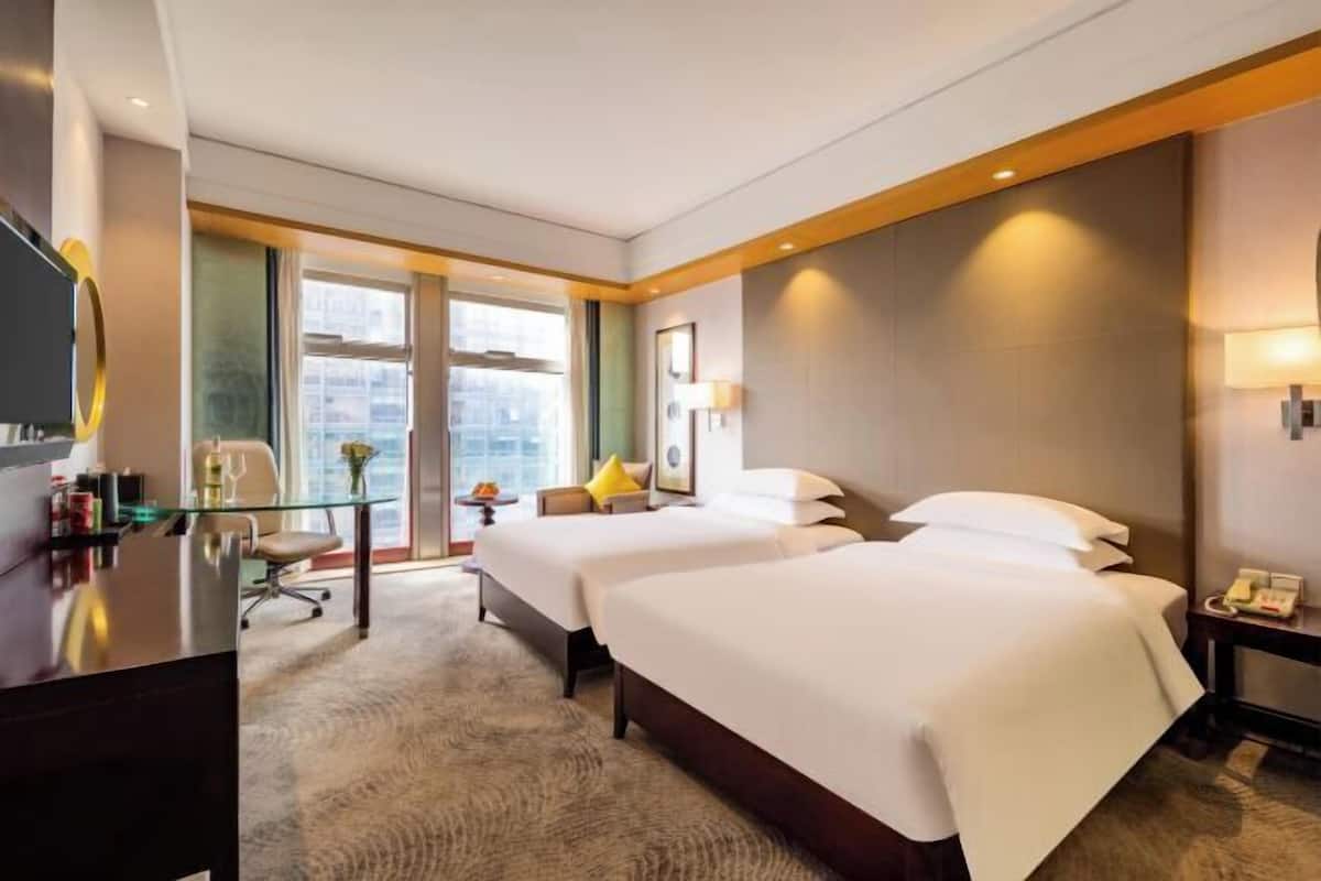 A hotel room with two beds, a desk, a chair, a TV, and a large window offering a city view.