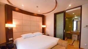 A hotel room with a large bed, two bedside lamps, a wooden headboard, a small seating area, and a bathroom visible through an open door.
