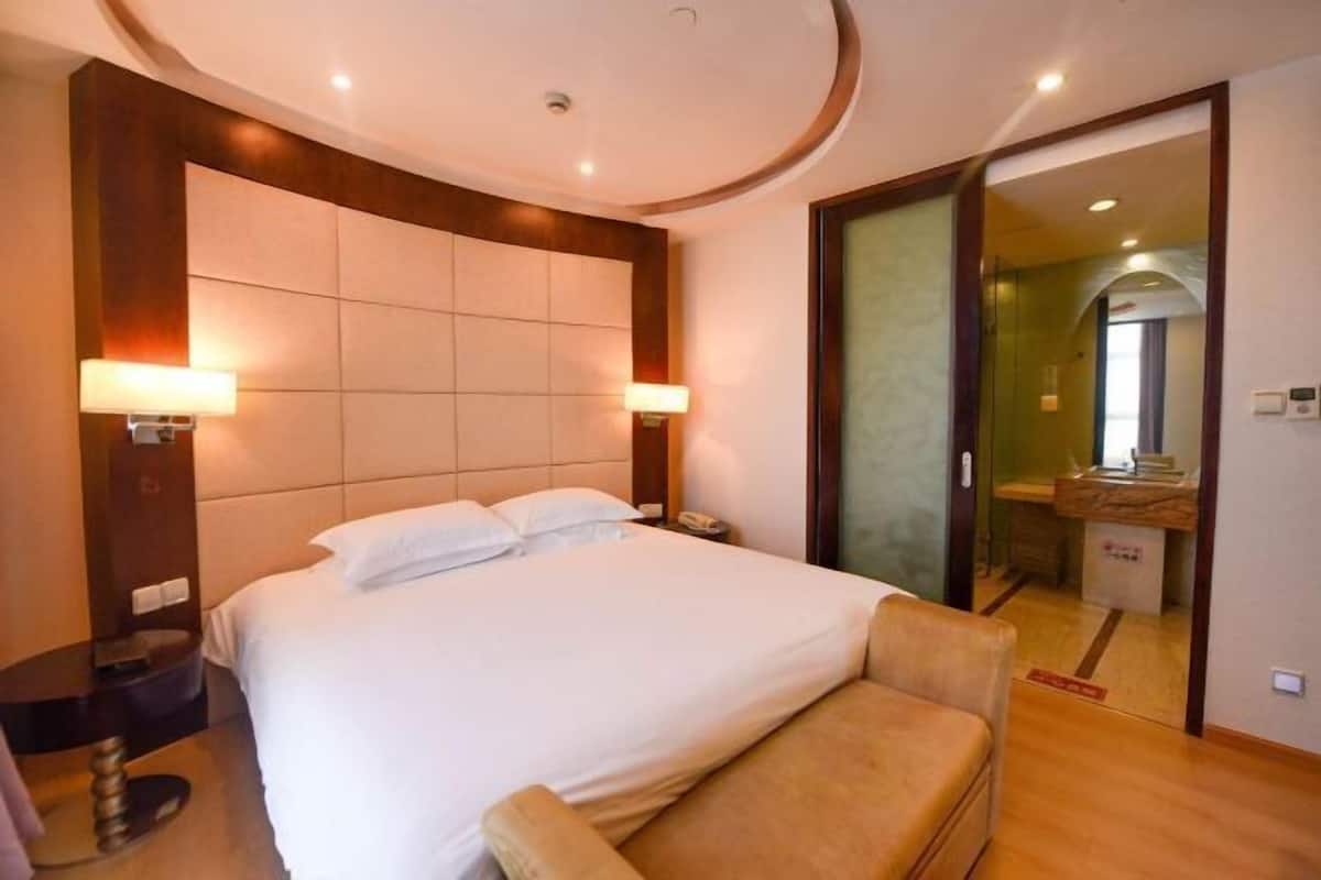 A hotel room with a large bed, two bedside lamps, a wooden headboard, a small seating area, and a bathroom visible through an open door.