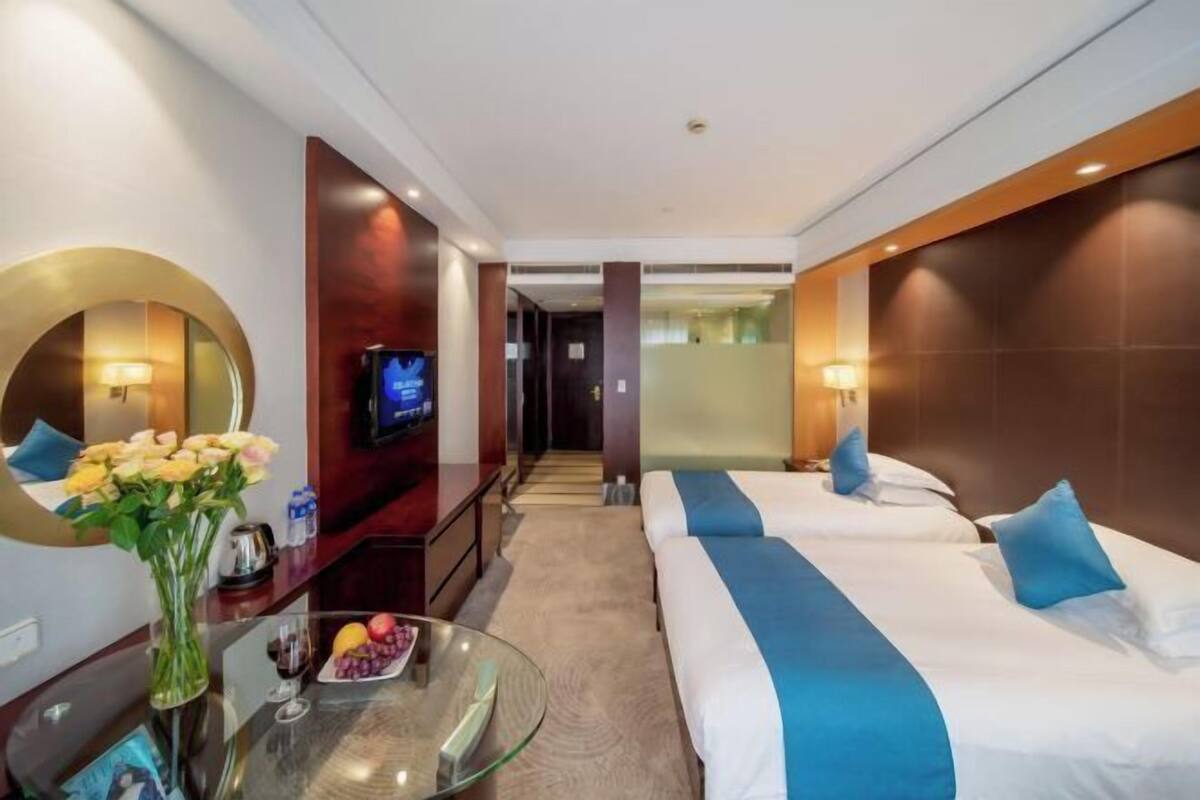 A hotel room with two beds, a TV, a glass table with a vase of flowers, and a view of the hallway.