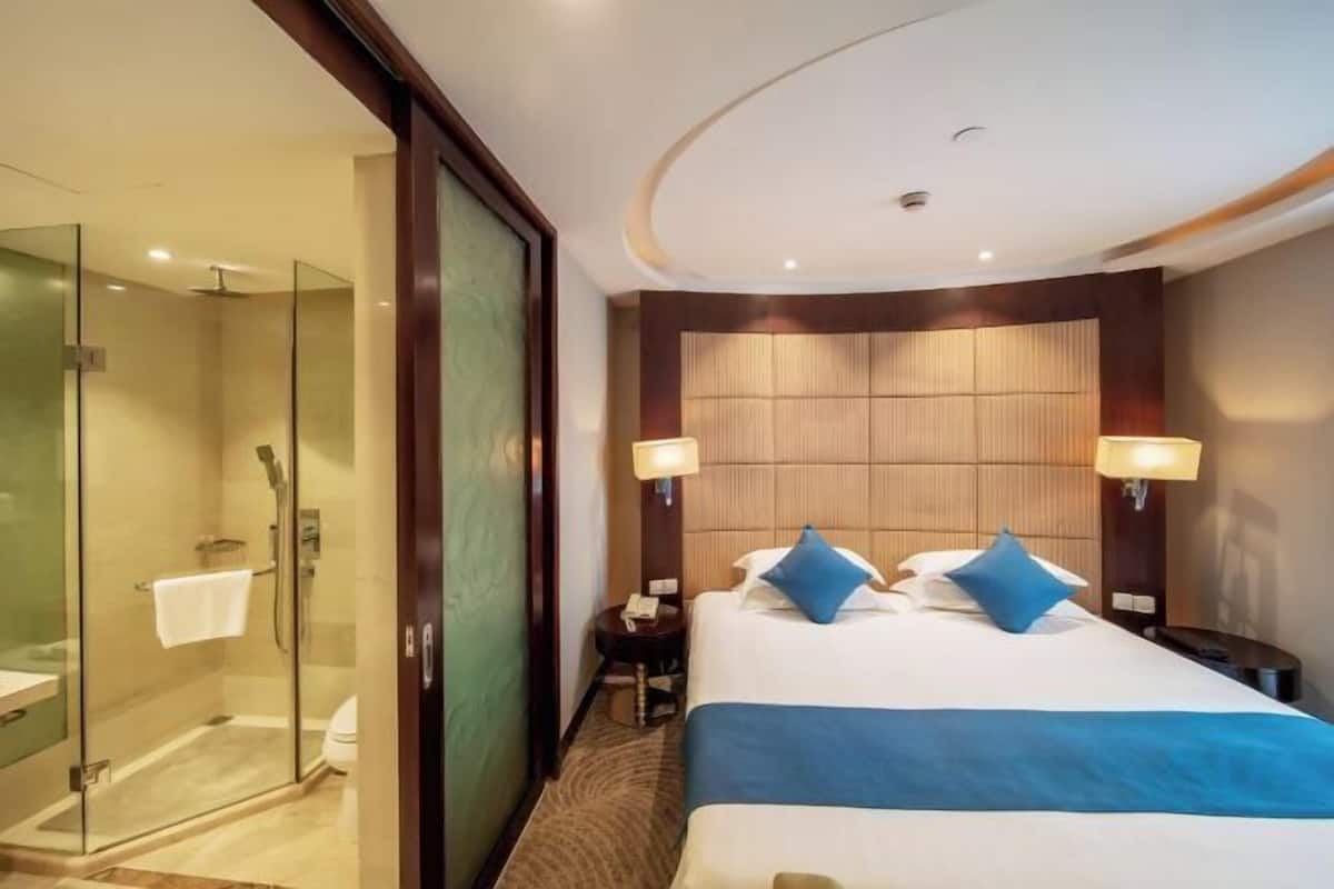 A hotel room with a glass-enclosed shower, a bed with blue bedding, and two bedside lamps.