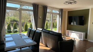 A modern living room with a dining area, large windows, a flat-screen TV, and a view of a garden.