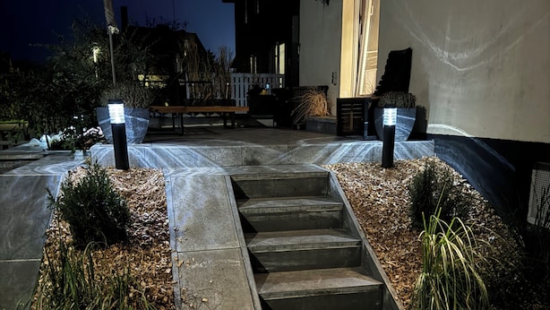 Un espace extérieur aménagé pour la nuit, avec un chemin en béton menant à l’entrée d’un bâtiment, entouré de parterres paysagers et éclairé par des luminaires encastrés dans le sol.
