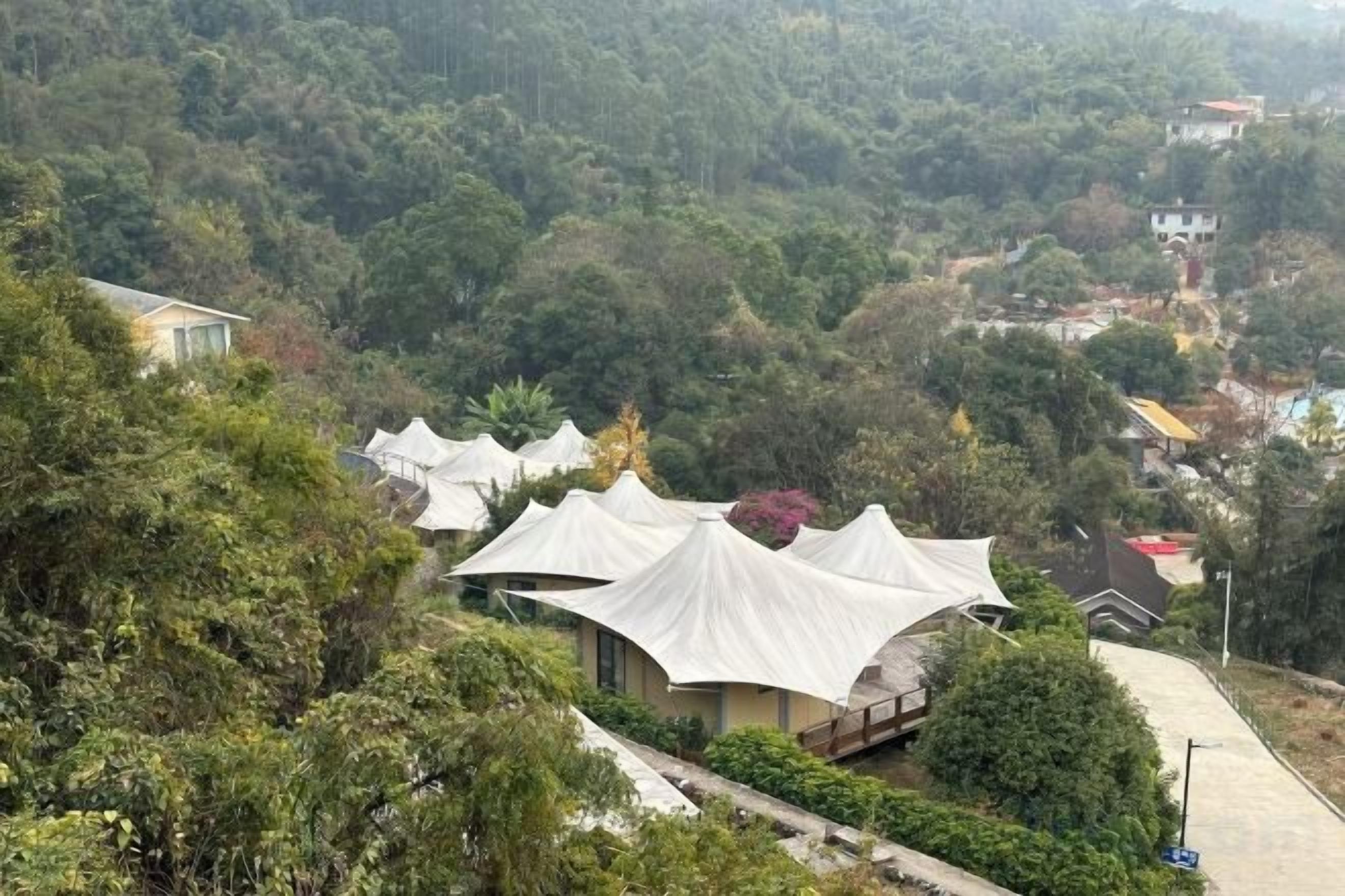 A series of white tents with a curved roof, surrounded by dense greenery and hills in the background.