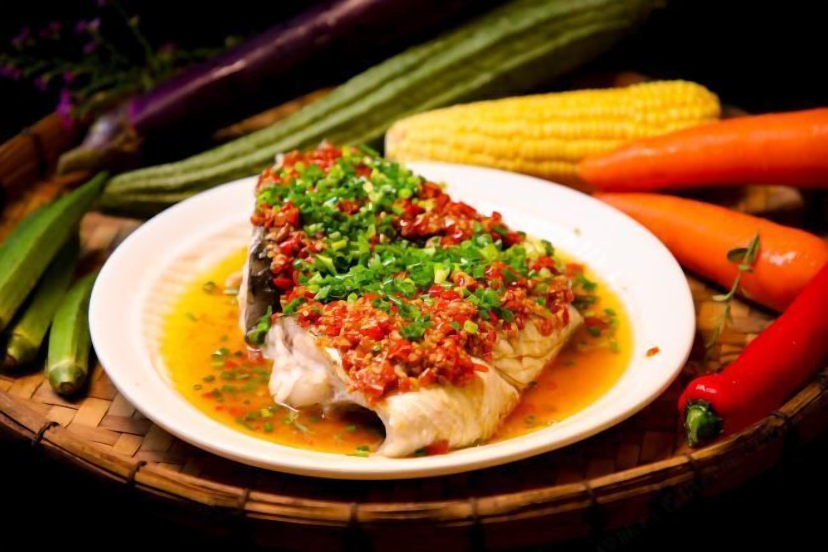 Une assiette de poisson nappée d’une sauce rouge et garnie d’herbes vertes, accompagnée de légumes comme du maïs et des carottes.