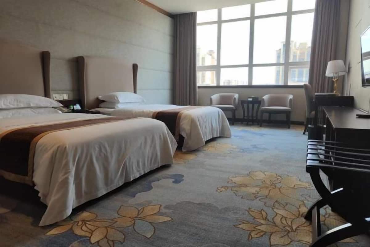 A hotel room with two beds, a desk, a chair, and a large window with a view of buildings.