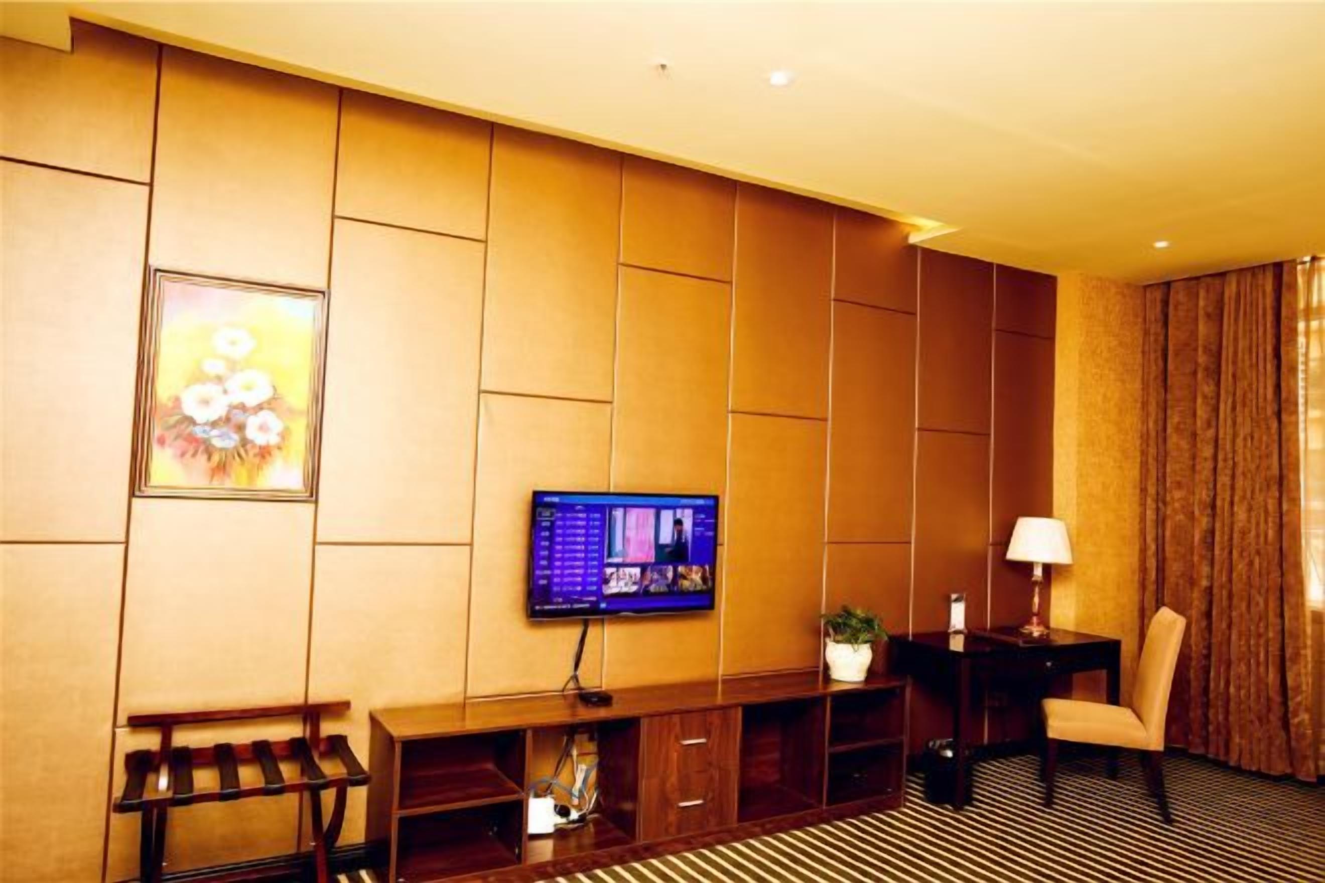 A hotel room with a flat-screen TV mounted on the wall, a small table with a lamp, a chair, and a bench with a wine rack.