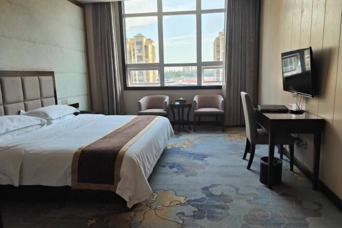 A hotel room with a bed, a desk, a chair, a television, and a large window with a view of buildings.