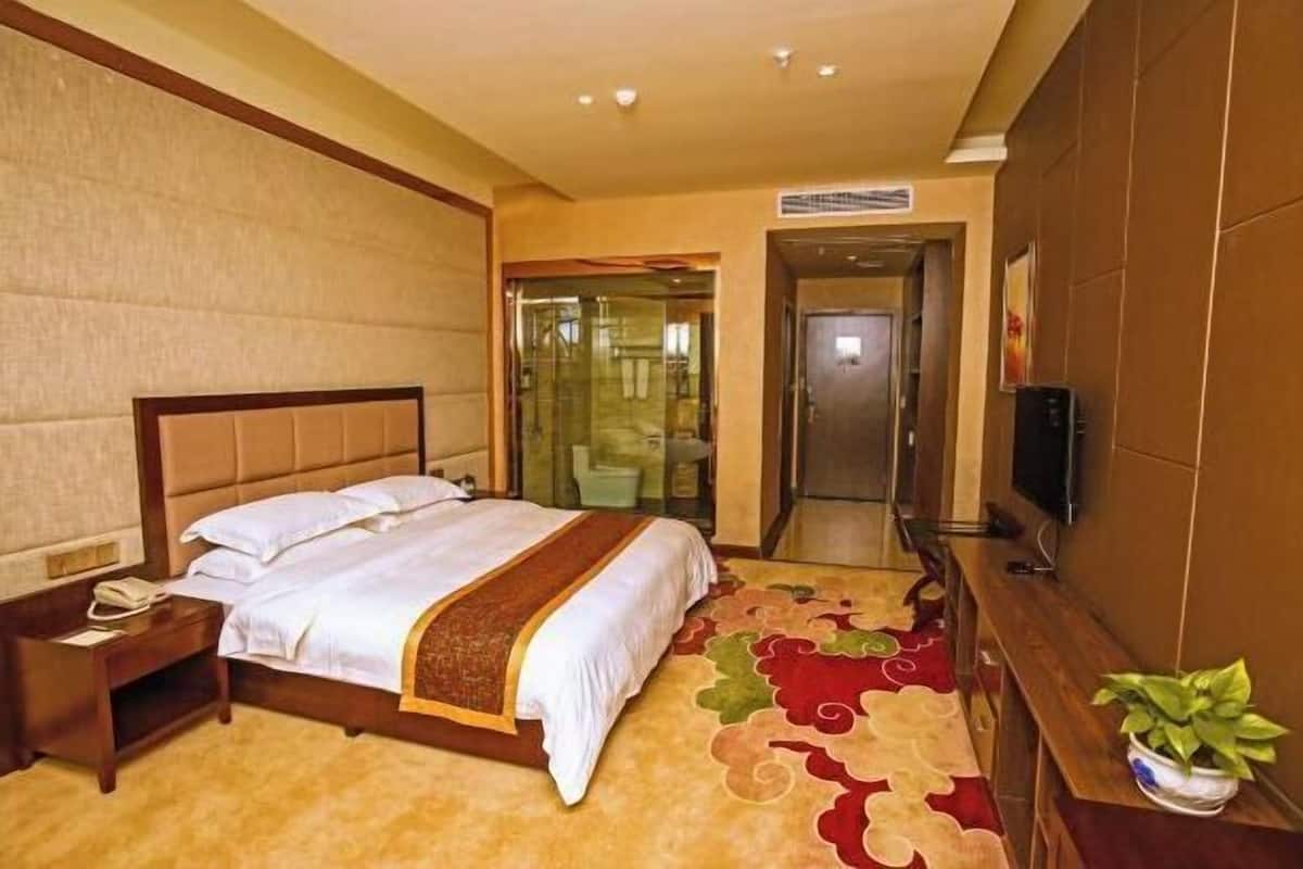 A hotel room with a bed, a nightstand, a television, and a bathroom visible through a glass door.