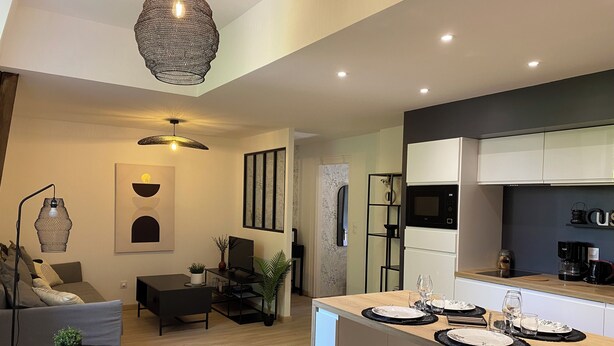 A modern kitchen with white cabinets, a black microwave, and a coffee machine. There is a dining area with a wooden table set for two, and a living room with a gray sofa and a black coffee table.