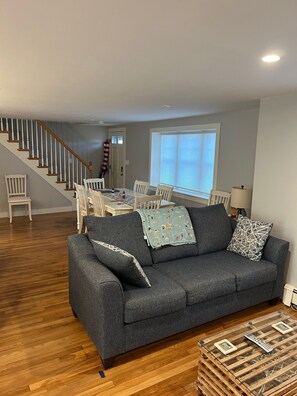 Ein Wohnzimmer mit grauem Sofa, Holzboden, einem Essbereich mit Tisch und Stühlen sowie einer Treppe.