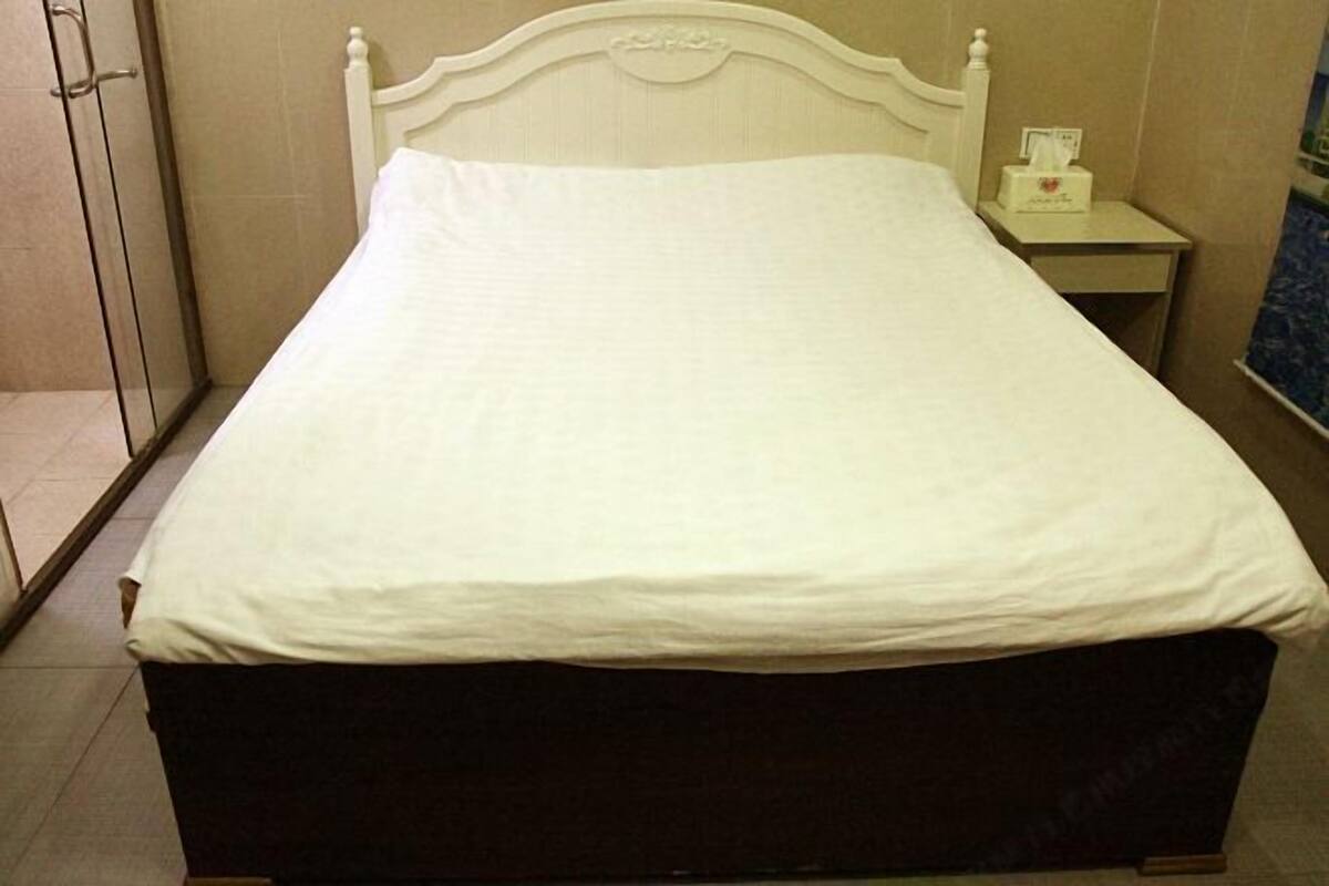 Un lit avec une tête de lit et un pied de lit blancs, une table de chevet avec une boîte de mouchoirs, et une zone douche située à gauche.