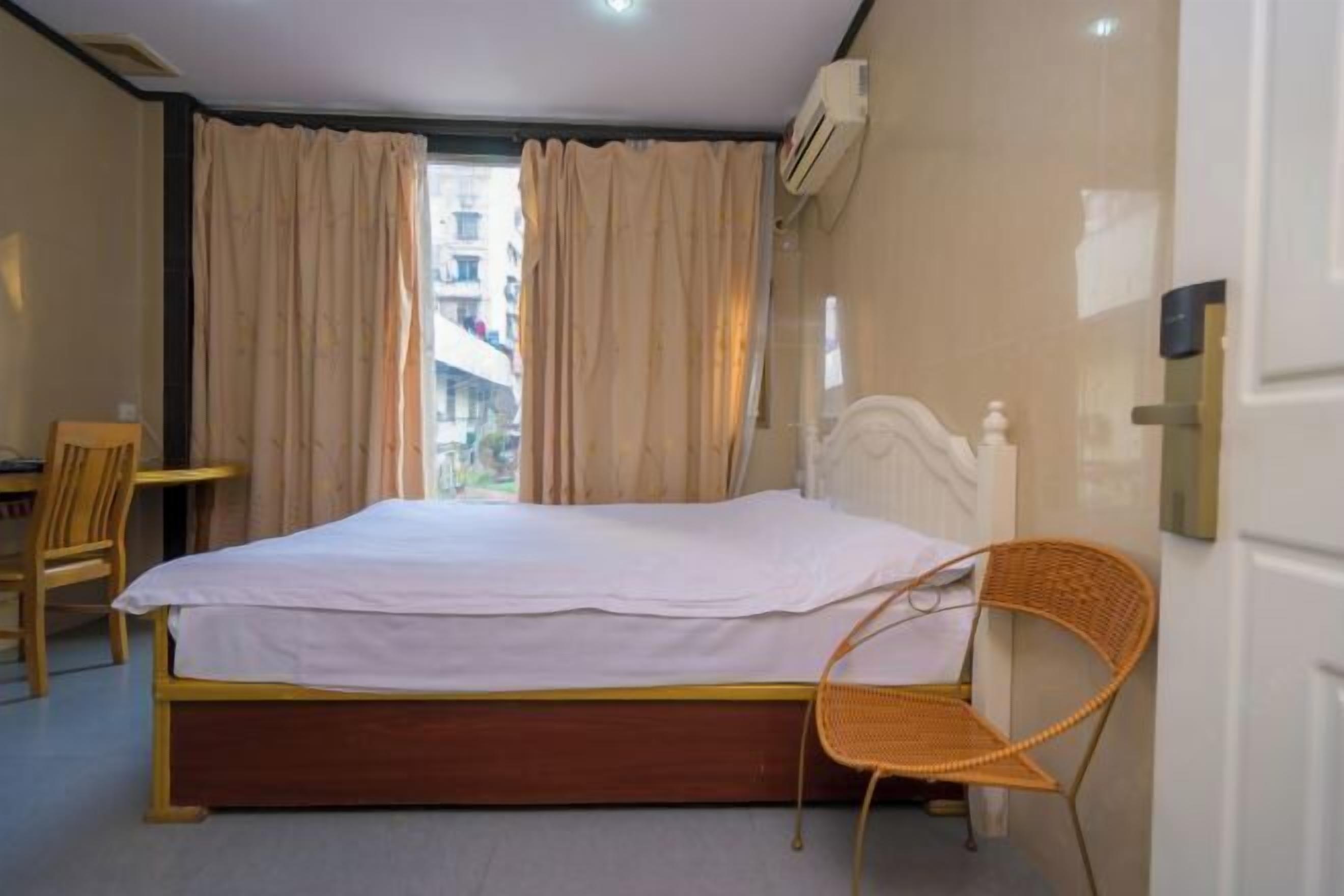Un dormitorio con cama, una silla, ventana con cortinas y unidad de aire acondicionado.