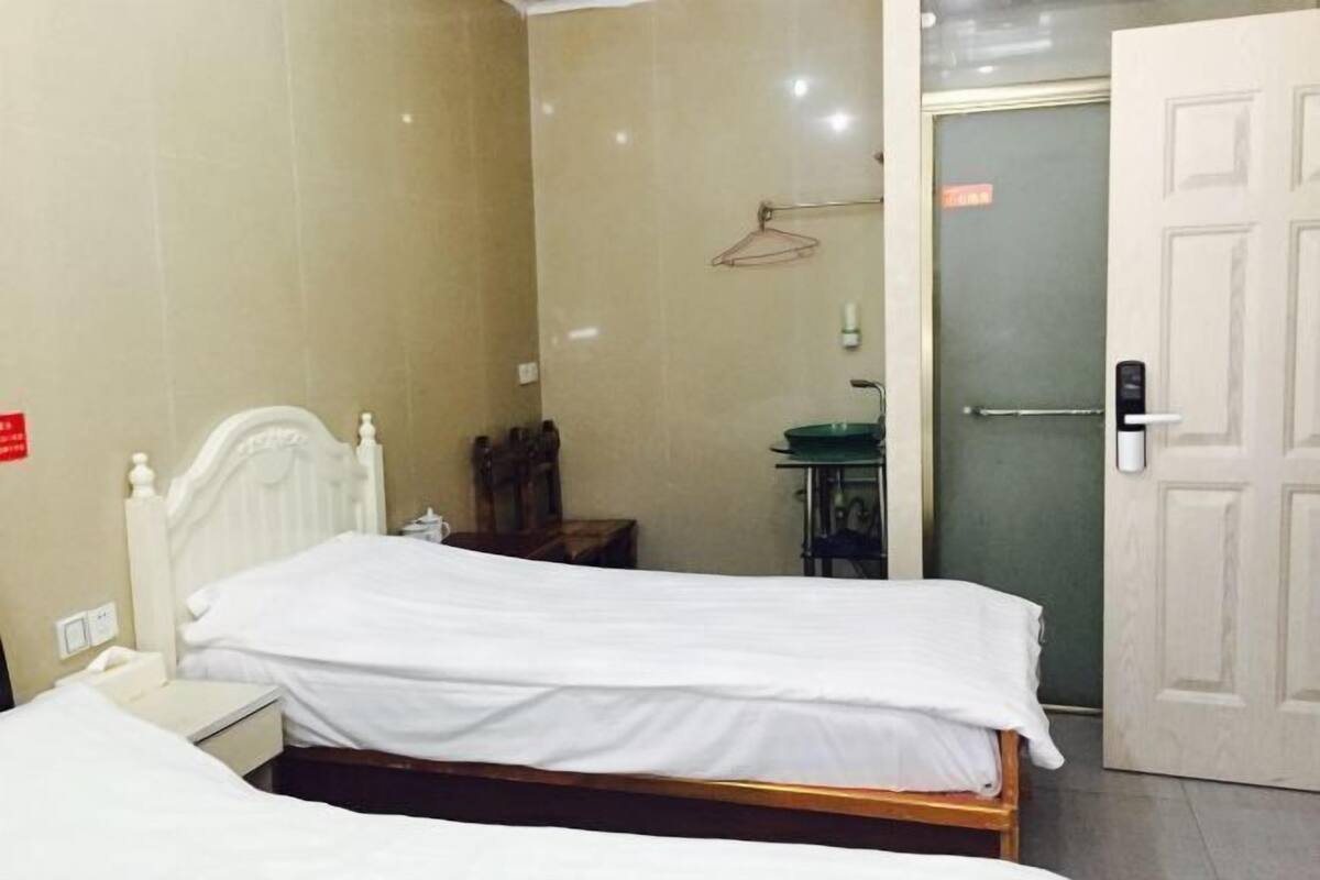 Une chambre d’hôtel avec deux lits, une table de chevet, une petite table, une chaise, une salle de bain avec un lavabo et un miroir, et une porte.