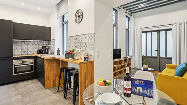 Una cocina moderna con armarios oscuros, una isla de madera y un comedor con un sofá amarillo y una mesa dispuesta para dos.