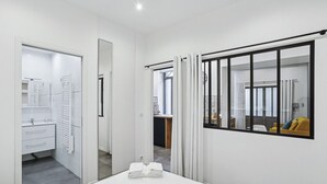 Habitación de hotel moderna con una cama, una mesita con toallas y un frutero, un baño con tendedero y vista a un salón a través de una ventana.