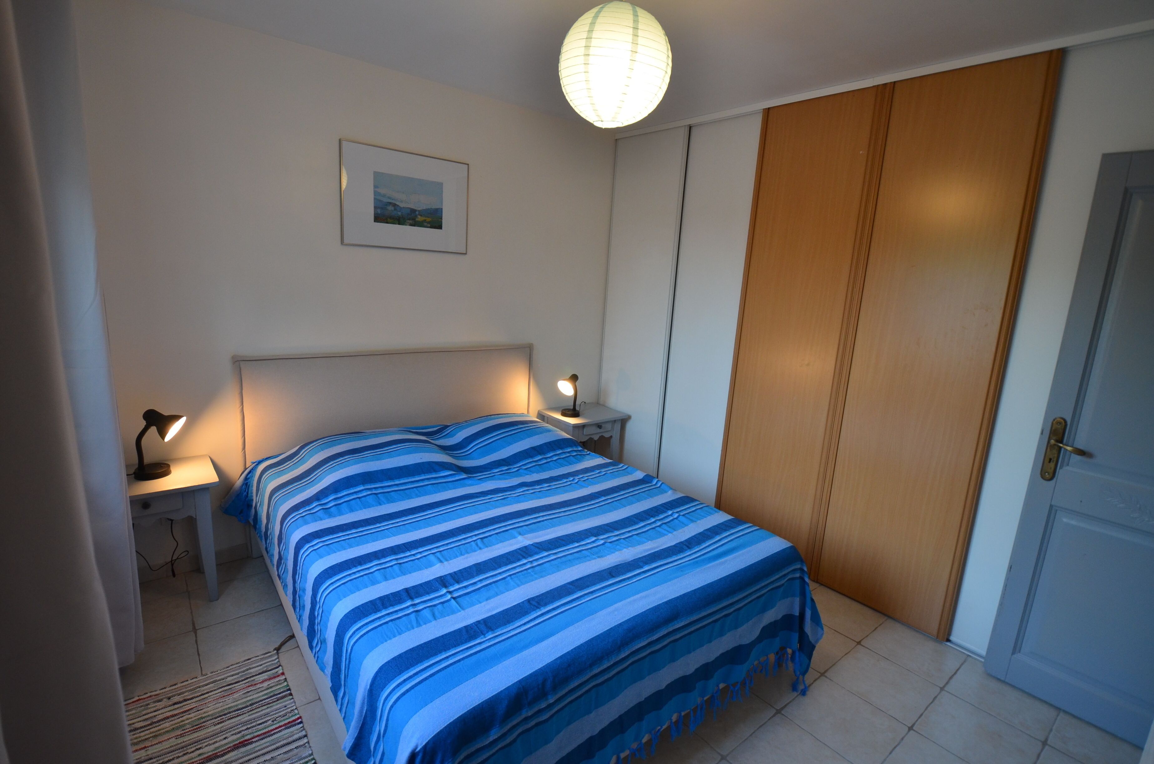 A bedroom with a bed, bedside tables, a lamp, a framed picture, a wardrobe, and a door.
