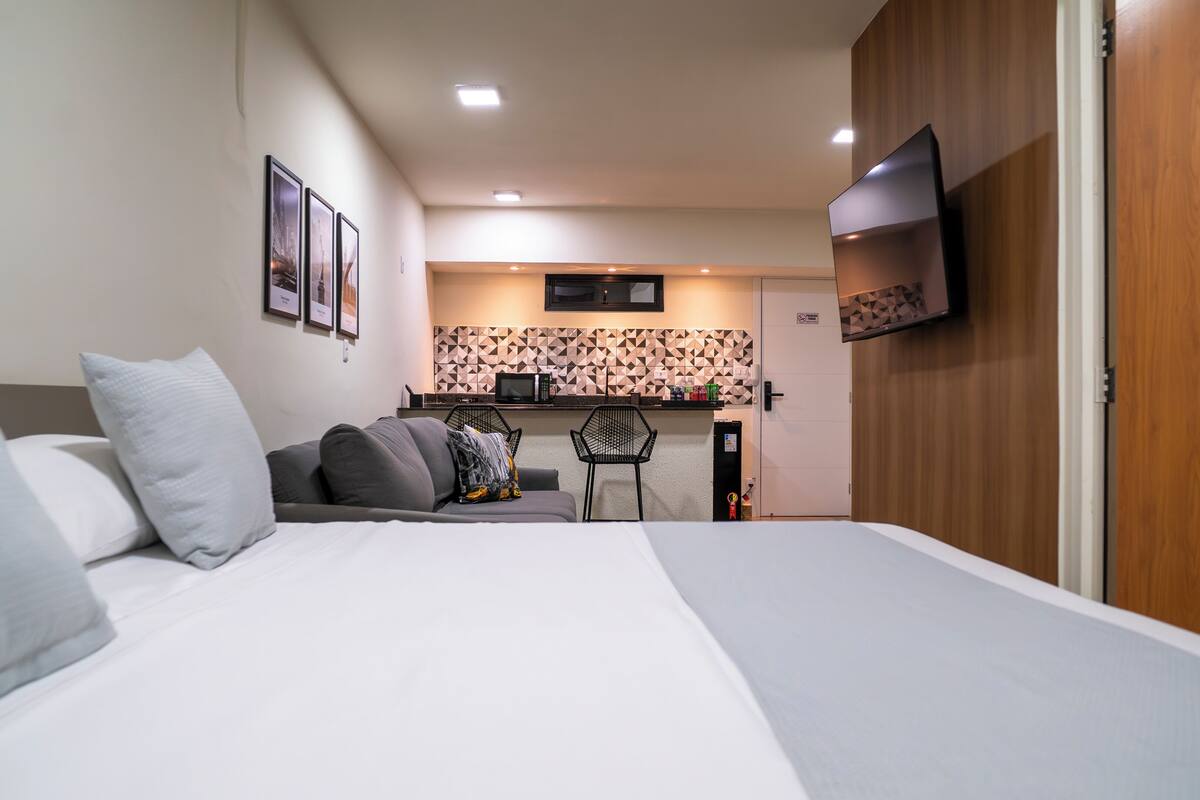 A bedroom with a bed, a sofa, a small kitchenette, and a TV mounted on the wall.