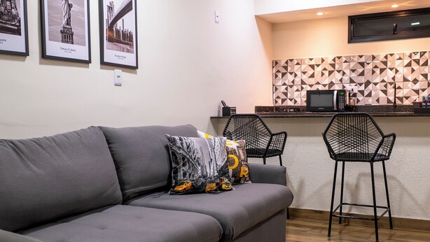 A modern living room with a grey sofa, two black chairs, and a kitchen area with a microwave and a geometric backsplash. - TNY APART BARRETOS (Barretos)
