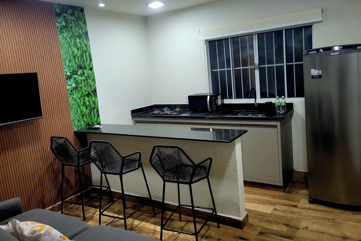 A modern kitchen with a central island, bar stools, a refrigerator, and a flat-screen TV mounted on the wall.