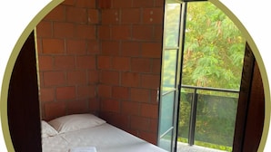 A single bed with a white bedspread and a folded towel in a room with brick walls and a glass door leading to an outdoor area with greenery. - Hotel Linda (Salto)