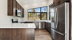A modern kitchen with dark wood cabinets, stainless steel appliances, and a large window offering a view of trees and a house. - Brand New, Luxury 1-bedroom apartment with AC, 10 Minutes to Downtown Whitefish (Whitefish)
