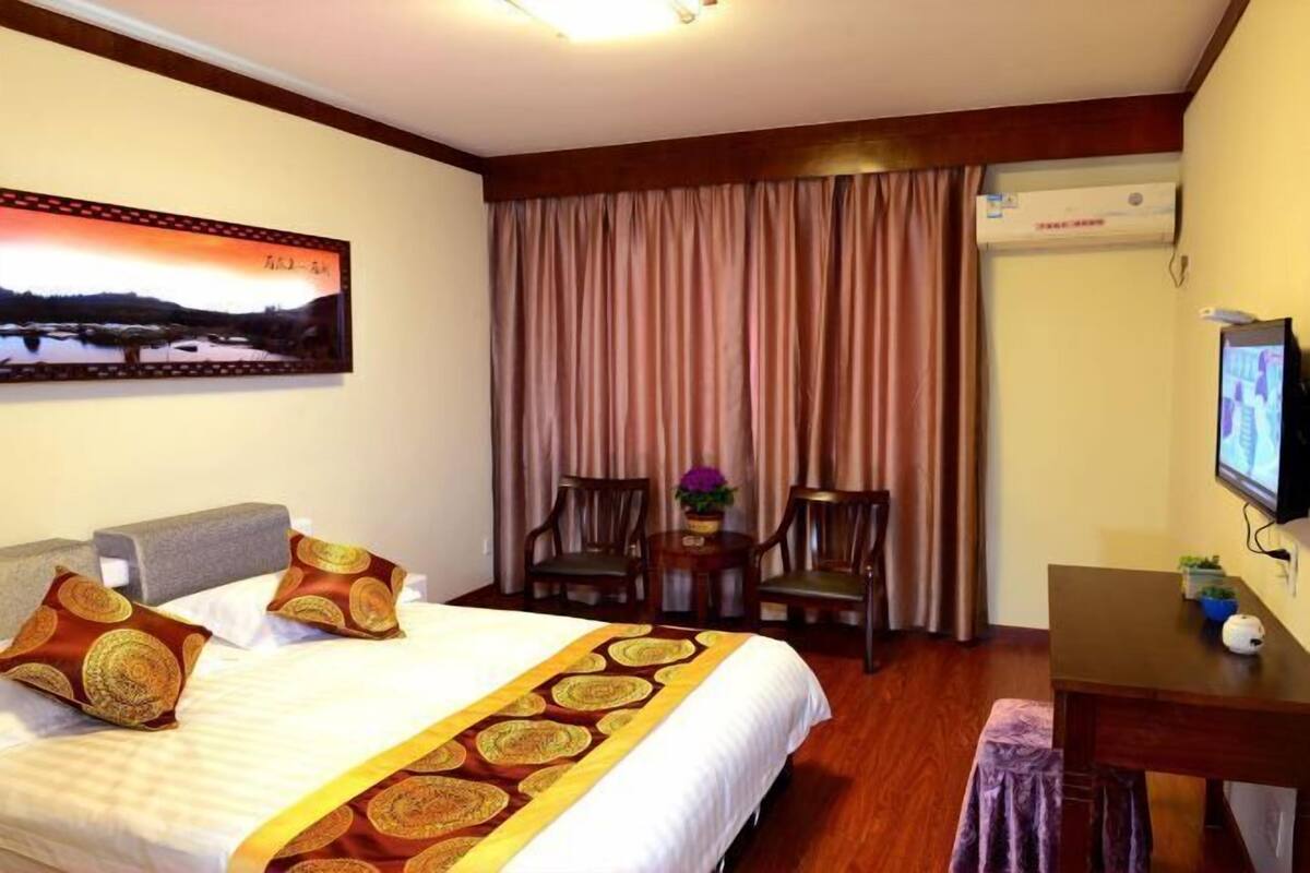 A hotel room with a bed, two chairs, a small table, a TV, and an air conditioning unit.