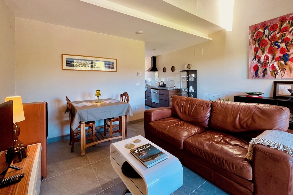 Una sala de estar con un sofá de cuero marrón, una mesa de comedor con sillas, una cocina al fondo y un cuadro en la pared.