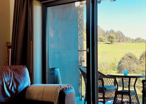 Een kamer met een schuifglazen deur die leidt naar een balkon met uitzicht op een groen veld en bomen.