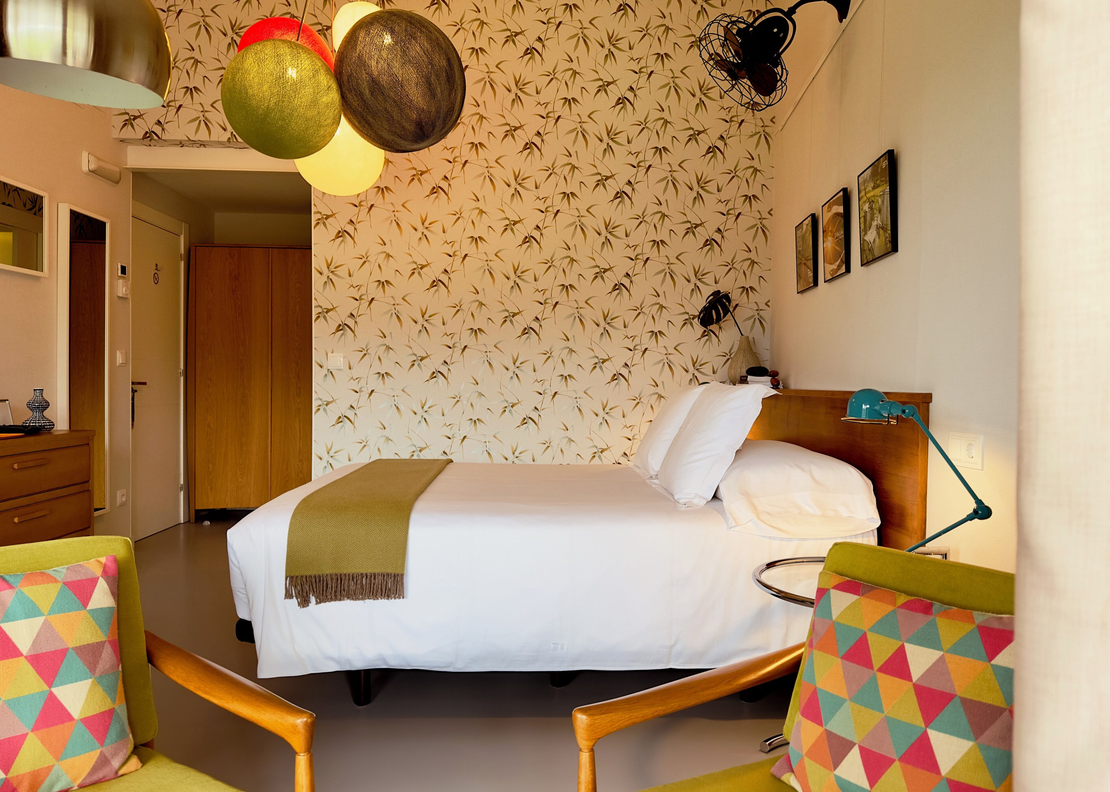 A hotel room with a bed, a bedside table with a lamp, a chair with a colorful cushion, and a wall with a bamboo pattern.