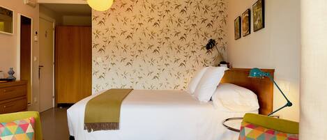 A hotel room with a bed, a bedside table with a lamp, a chair with a colorful cushion, and a wall with a bamboo pattern.