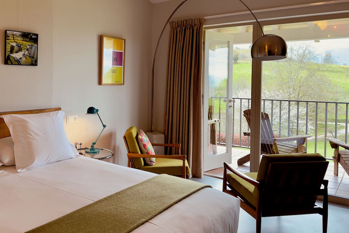 Habitación de hotel con una cama grande, una silla, un escritorio, una lámpara y un balcón con vistas al exterior.