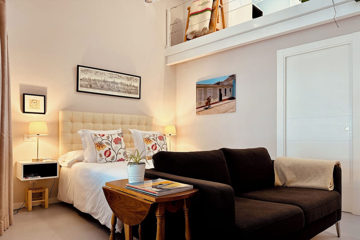 Un dormitorio con cama, mesita de noche, una mesa con libros, planta en maceta, sofá y dos cuadros colgados en la pared.