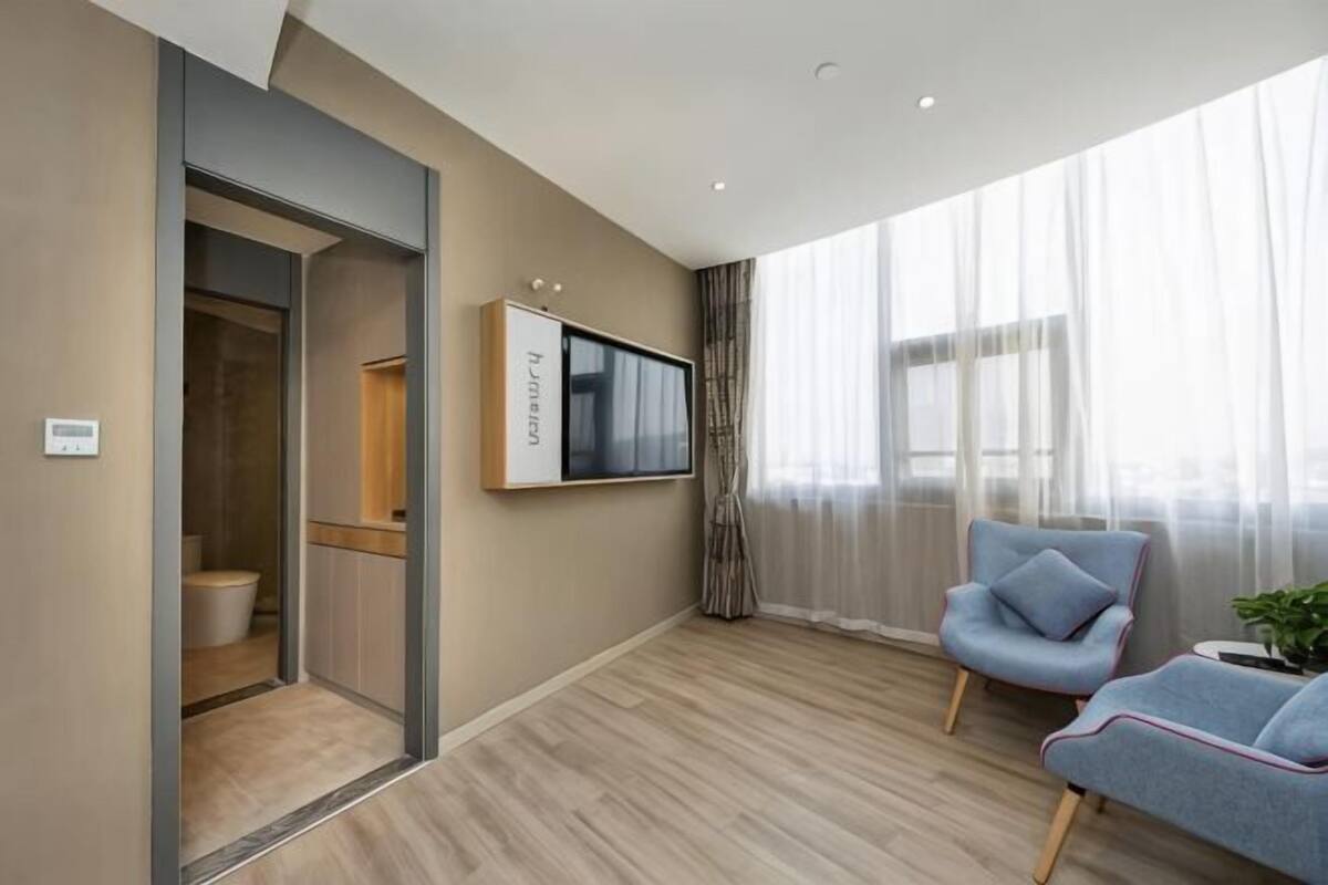 Moderni hotellihuone, jossa on suuri ikkuna, seinälle asennettu taulutelevisio ja oleskelualue, jossa on sininen nojatuoli.