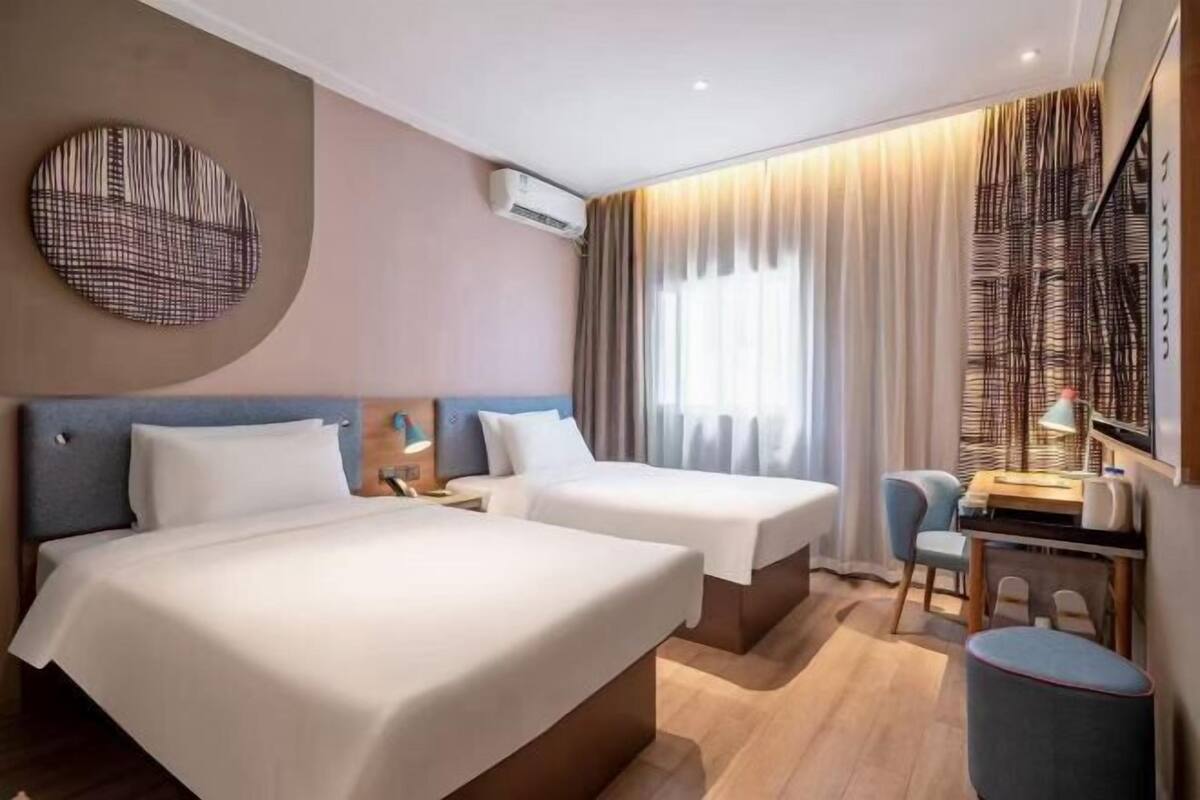Hotellihuone, jossa on kaksi sänkyä, työpöytä, tuoli ja suuri ikkuna, jossa on verhot.