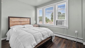 Un dormitorio con cama de tablero de madera, ropa de cama blanca y una ventana con persianas.