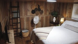 Une pièce chaleureuse avec un mur en bois, un lit recouvert d’une couverture à rayures, un ventilateur sur pied, une petite plante et une échelle.