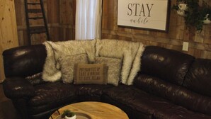 Un canapé d’angle en cuir marron foncé est agrémenté d’un plaid blanc moelleux et d’un coussin sur lequel on peut lire : « RELÂCHEZ-VOUS, LEVEZ LES PIEDS, ASSEZ-VOUS ET DÉTENDEZ-VOUS ». Une petite table ronde en bois, sur laquelle est posée une plante, se trouve devant le canapé. Une échelle est appuyée contre le mur, à gauche, et une pancarte au-dessus du canapé indique : « RESTEZ UN PEU ».