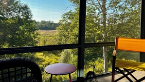Een kamer met een groot raam dat uitkijkt op een groen landschap, een klein rond tafeltje en twee stoelen.
