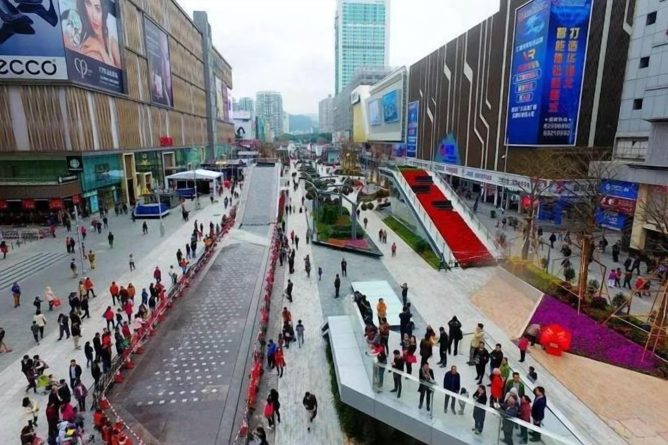 A busy urban square with a central pedestrian area, surrounded by modern buildings and advertisements.