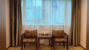 Une pièce avec deux chaises en bois et une petite table, une fenêtre ornée de rideaux légers, et une vue sur les bâtiments à l’extérieur.