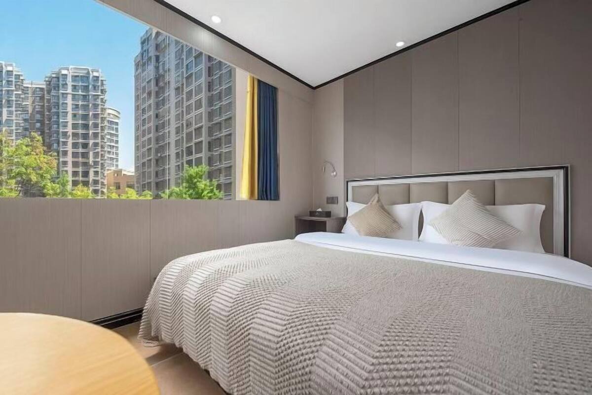 Une chambre moderne avec un grand lit, une table de chevet et une fenêtre donnant sur des immeubles de grande hauteur.