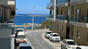A street view with parked cars and a building with balconies overlooking a coastal area.