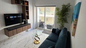 A modern living room with a flat-screen TV, a dark sofa, a glass coffee table, and a large window with a view of buildings.