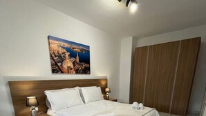 A hotel room with a large bed, a nightstand with a lamp, a wooden headboard, a painting of a cityscape, and a wardrobe.