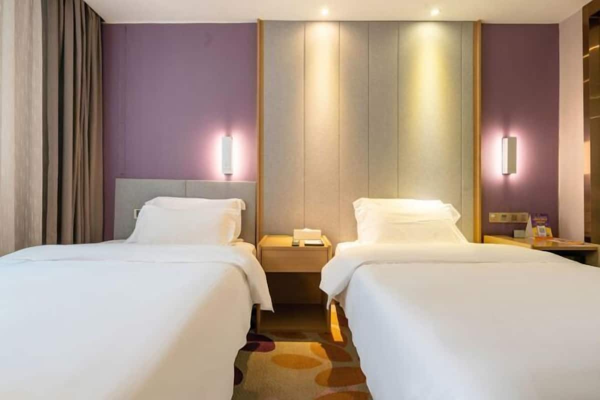 Hotellihuone, jossa on kaksi sänkyä, y öpöytä, seinään kiinnitetty lamppu ja matolla peitetty lattia.