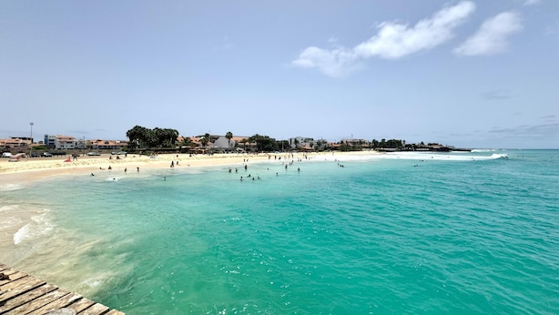 Ein Strand mit klarem türkisblauem Wasser, einer Holzterrasse und Gebäuden im Hintergrund.