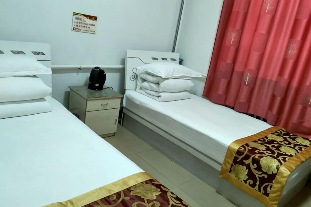 A hotel room with two single beds, a nightstand with a kettle, and a red curtain.