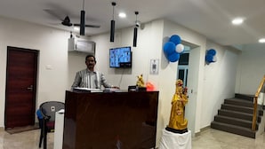 A reception area with a marble reception desk, a ceiling fan, a mounted television, and a statue of a religious figure. - L D Residency (Kilvelur)