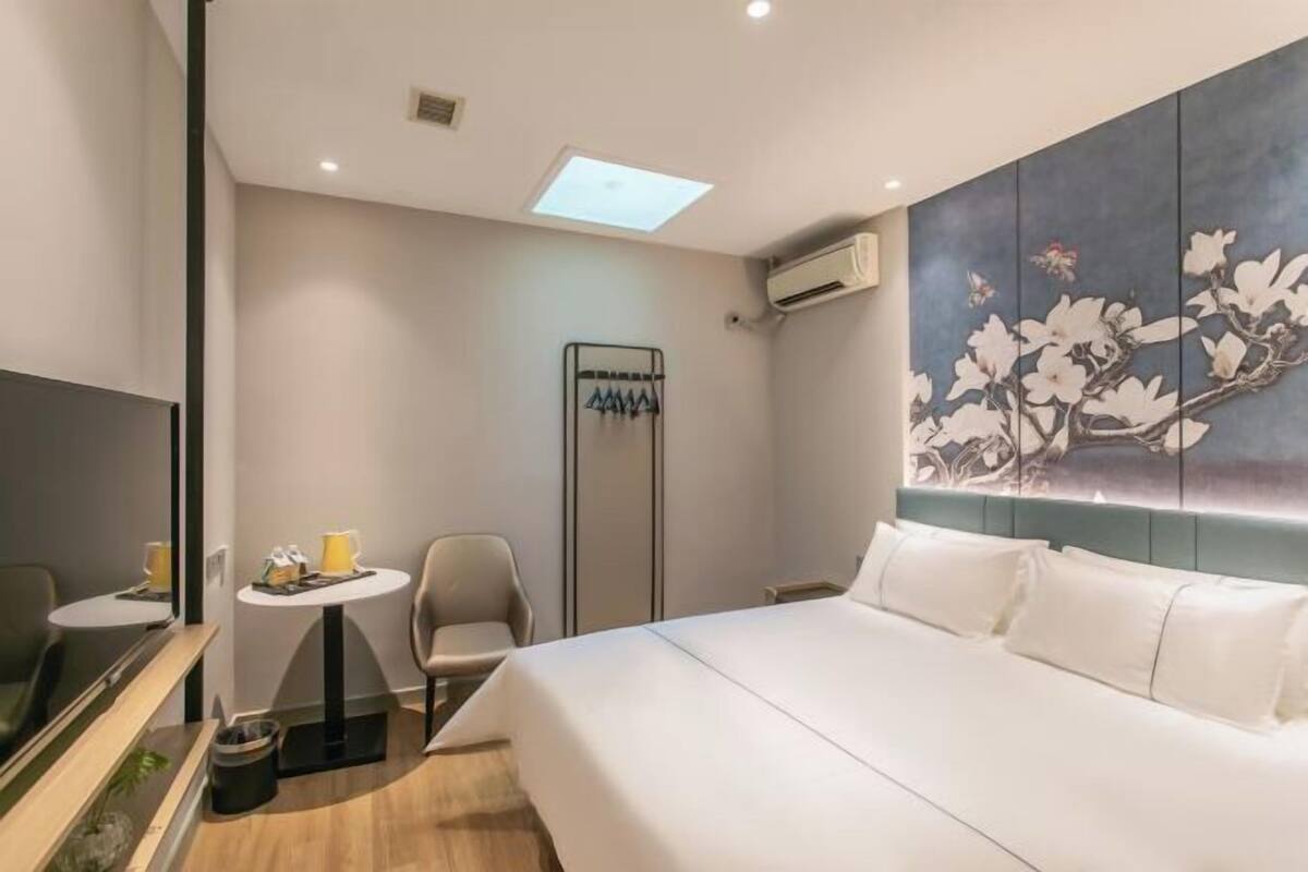 Una habitación de hotel con cama, una mesita, una silla, televisor y aire acondicionado instalado en la pared.